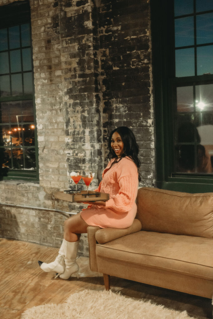 Sharece, owner of Milwaukee business On the Rocks Mobile Bartending Company, sitting on the arm of a leather sofa, smiling, holding a wooden tray of pink cosmos with a round gold branded drink topper with their logo on the rim and a white rose in the glass