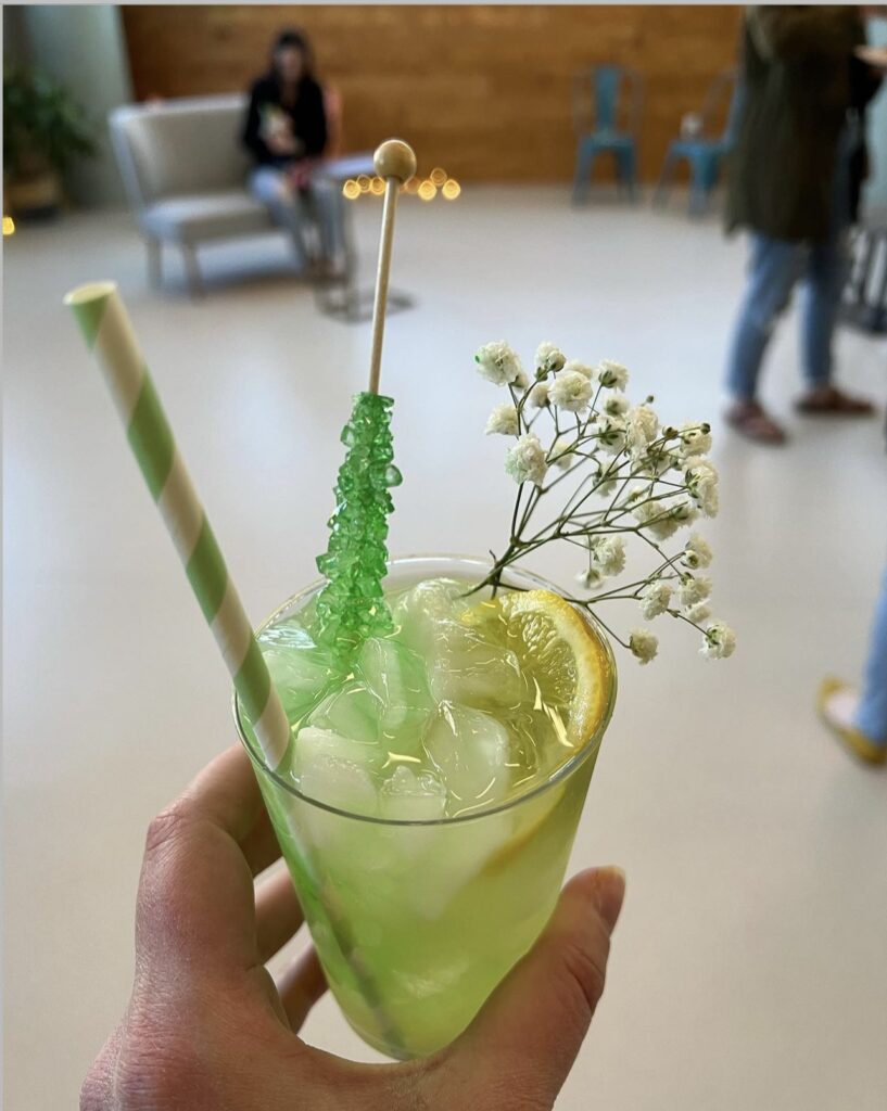 kiwi lemonade mocktail in a clear glass with lemon slice garnishes as well as baby’s breath and green rock candy sticks resting on the rim