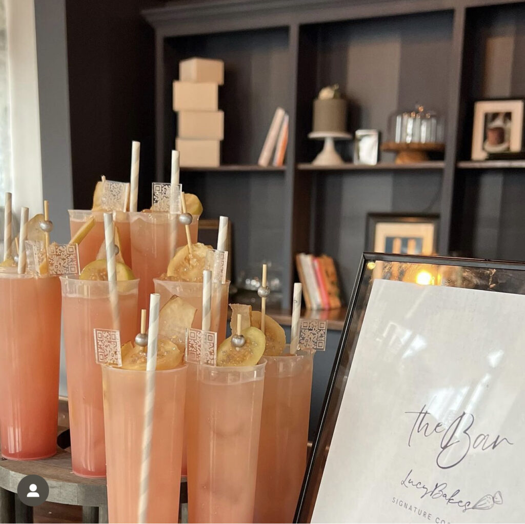 A custom bar and bartending setup by On the Rocks Mobile Bartending Company for a grand opening event for a local Milwaukee business, closeup of the drink display where the drinks are displayed at 3 different heights forming a beautiful look, the drinks are in tall clear glasses and are a pink cocktail with fresh lemon slice garnishes lining a stir stick with a grey pearl on the end, a grey and white paper stray and a clear acrylic drink stirrer with a QR code at the top with the business’s website