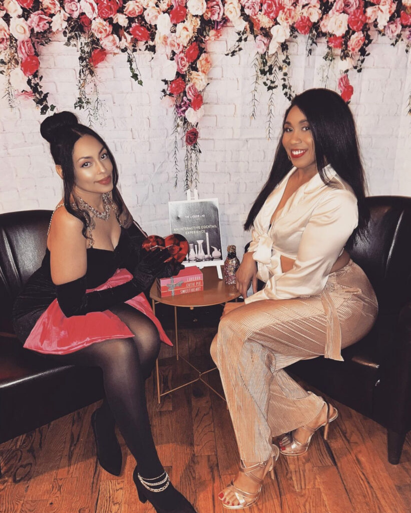 The Liquor Lab founders Courtney and Sharece at the Valentine’s Day Liquor Lab Cocktail Making Experience Pop-Up in Milwaukee, WI sitting on leather chairs with a backdrop of hanging white, pink and red roses