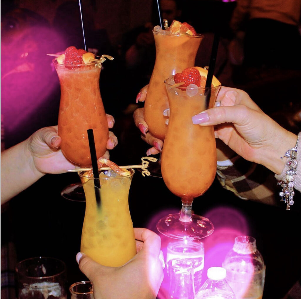 4 people’s hands cheersing their cocktails created at The Liquor Lab, an interactive cocktail making experience pop-up event in Milwaukee, WI. The drinks are vibrant orange and yellow with fresh strawberry and orange slice garnishes and a drink stirrer that says “love” In cursive for their Valentine’s Day themed event