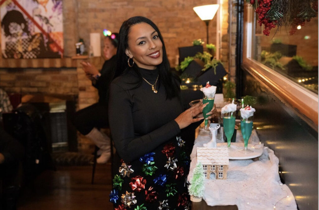 Liquor Lab Christmas holiday event cocktail-making pop up event in Milwaukee, WI with founder Courtney smiling at camera holding a green cocktail with whipped topping and red and white Hershey kiss topper and green and white straw, over the holiday display with drinks, snow and twinkle light and gingerbread display