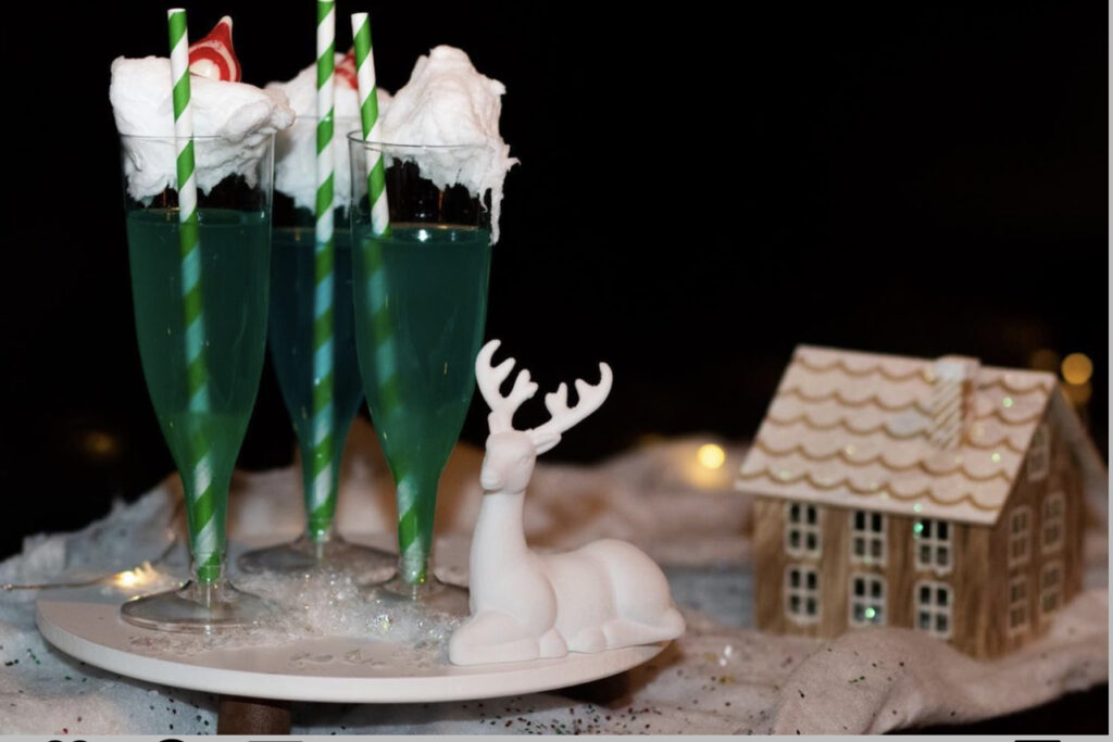 Liquor Lab Christmas holiday event cocktail-making pop up event in Milwaukee, WI bar display with a white tray of green cocktail with whipped topping and red and white Hershey kiss topper and green and white straw, over the holiday display with drinks, snow and twinkle light and gingerbread display