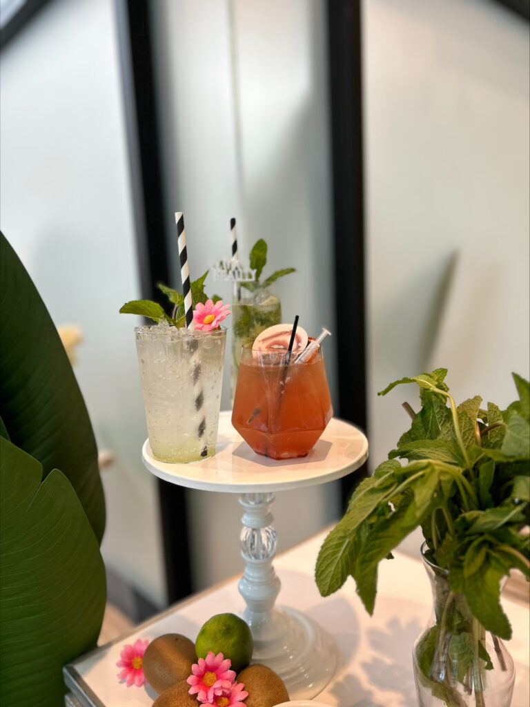 corporate event bar setup by On the Rocks Mobile Bartending Company for a Madison, WI business the drinks displayed on a cute white cake stand include a cocktail with a round paper drink topper that matches the theme of the event, a clear refreshing mojito drink with a black and white striped paper straw, fresh mint garnish and a small pink flower, and the extra garnishes are in small white bowls