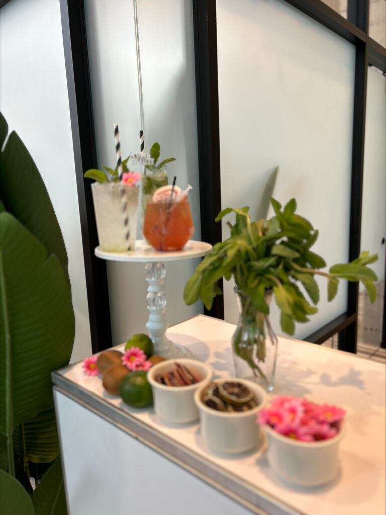 corporate event bar setup by On the Rocks Mobile Bartending Company for a Madison, WI business the drinks displayed on a cute white cake stand include a cocktail with a round paper drink topper that matches the theme of the event, a clear refreshing mojito drink with a black and white striped paper straw, fresh mint garnish and a small pink flower, and the extra garnishes are in small white bowls