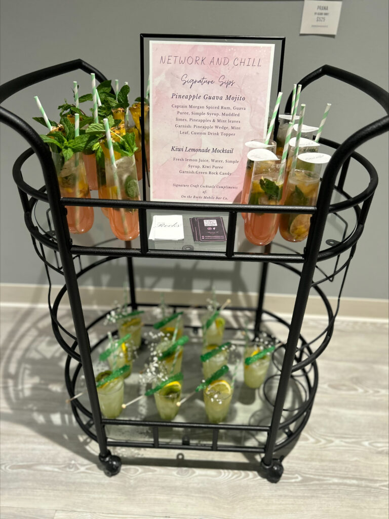 Bar setup and cocktail/mocktail display for a Milwaukee networking event designed by On the Rocks Mobile Bartending Company. The drinks are on a black multi-level wire and clear shelf with a pastel pink watercolor menu sign. The drinks on the top shelf are a pineapple guava mojito with fresh mint and lime slice garnishes, a green and yellow striped paper straw and a paper drink topper with the logo of the event. The bottom shelf drinks are a kiwi lemonade mocktail in a clear glass with lemon slice garnishes as well as baby’s breath and green rock candy sticks resting on the rim