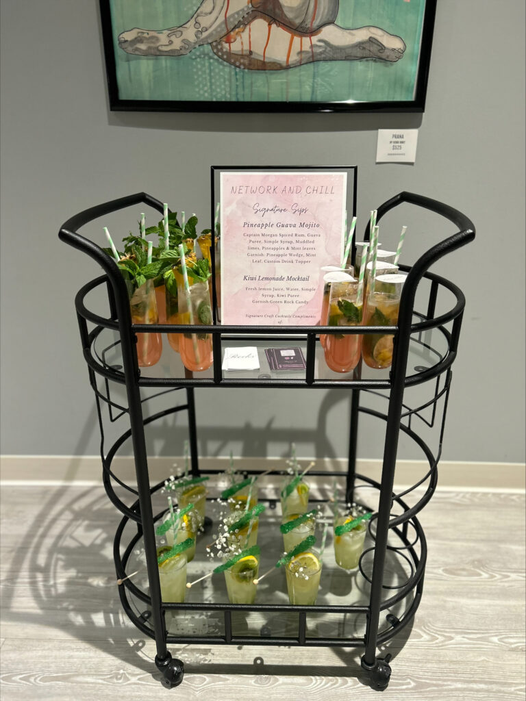 Bar setup and cocktail/mocktail display for a Milwaukee networking event designed by On the Rocks Mobile Bartending Company. The drinks are on a black multi-level wire and clear shelf with a pastel pink watercolor menu sign. The drinks on the top shelf are a pineapple guava mojito with fresh mint and lime slice garnishes, a green and yellow striped paper straw and a paper drink topper with the logo of the event. The bottom shelf drinks are a kiwi lemonade mocktail in a clear glass with lemon slice garnishes as well as baby’s breath and green rock candy sticks resting on the rim