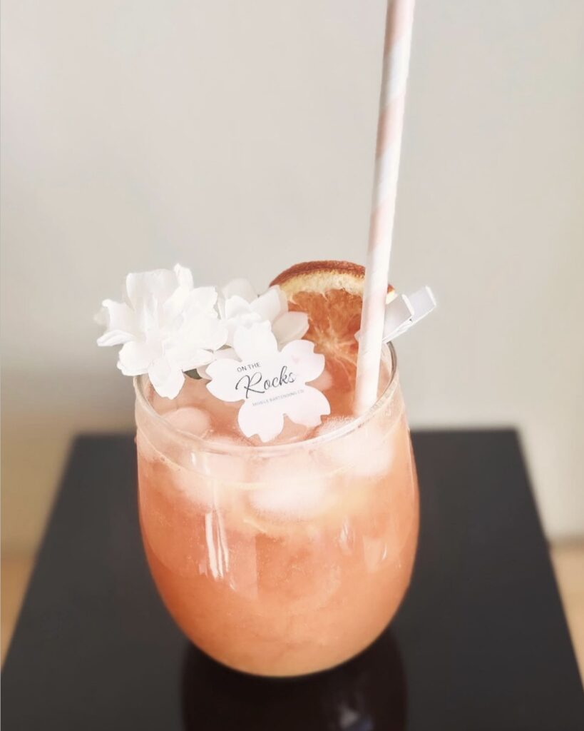 Close up view looking straight on of on a beautiful cocktail created by On the Rocks Mobile Bartending Company in Milwaukee, WI of a round clear glass with a pink drink, round ice cubes, a pastel pink and white paper straw, fresh orange slice garnish pinned to the rim with a small white clothespin, cute little white flowers topping the drink and a paper drink topper in the shape of a flower with On the Rocks’ logo displayed on it