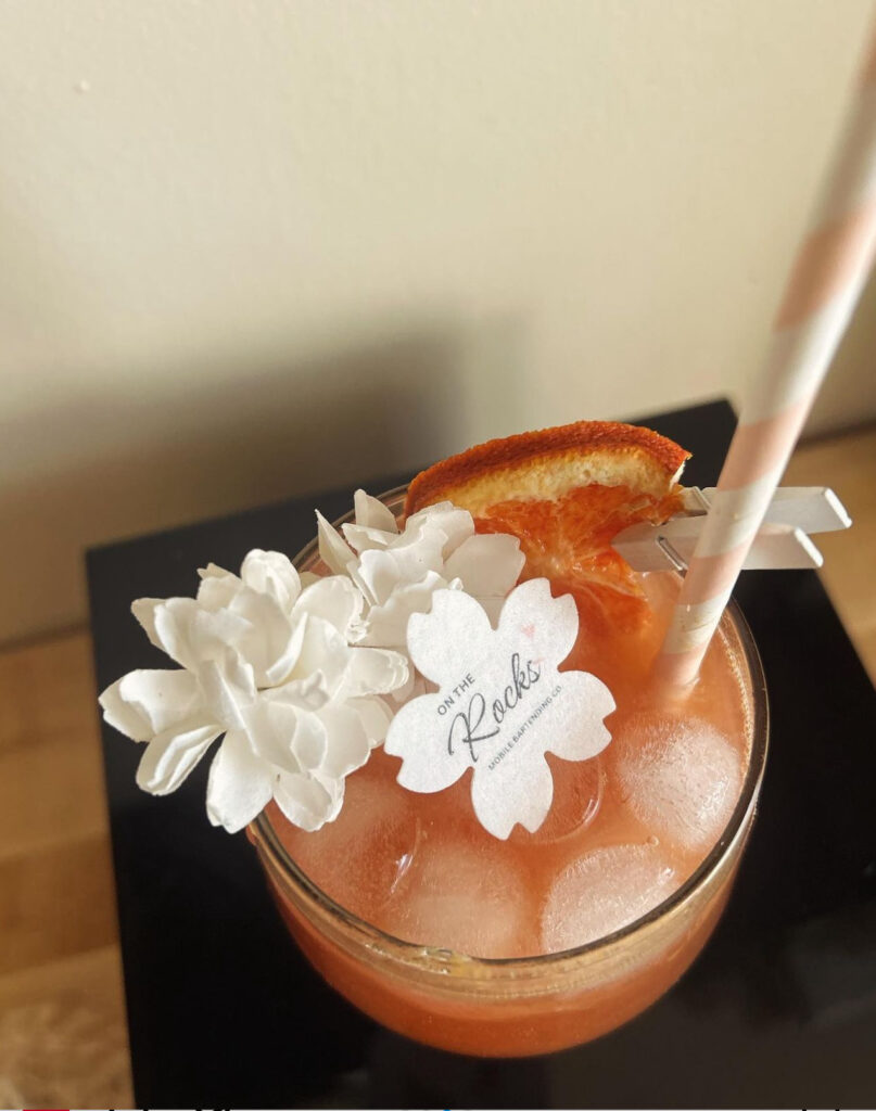 Close up view looking down on a beautiful cocktail created by On the Rocks Mobile Bartending Company in Milwaukee, WI of a round clear glass with a pink drink, round ice cubes, a pastel pink and white paper straw, fresh orange slice garnish pinned to the rim with a small white clothespin, cute little white flowers topping the drink and a paper drink topper in the shape of a flower with On the Rocks’ logo displayed on it