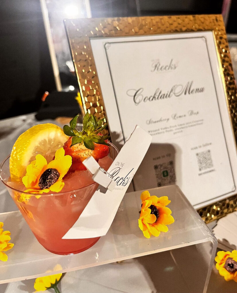 Display of a bar setup by On the Rocks Mobile Bartending Co for a Milwaukee event with a vintage thick gold framed cocktail menu and a beautiful vibrant pink drink in a clear glass with fresh strawberry and lemon slice garnishes as well as a pretty yellow flower topping the drink. Pretty yellow flowers are decorating the bar display for extra wow factor making it a beautiful bar display matching the theme of the event