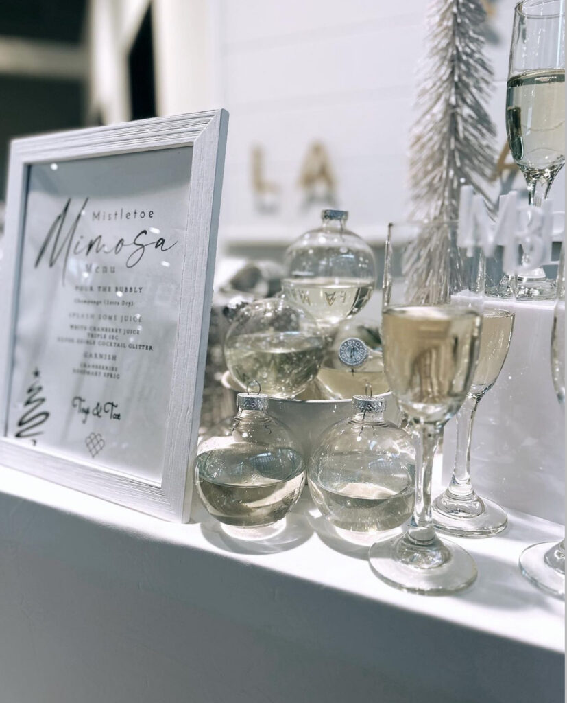 Stunning holiday corporate event mobile bartending bar setup in Madison, WI of white table, mimosa bar, shiny silver holiday décor, champagne glasses and holiday clear ball ornaments filled with champagne for a gorgeous design touch next to a white framed mistletoe mimosa menu