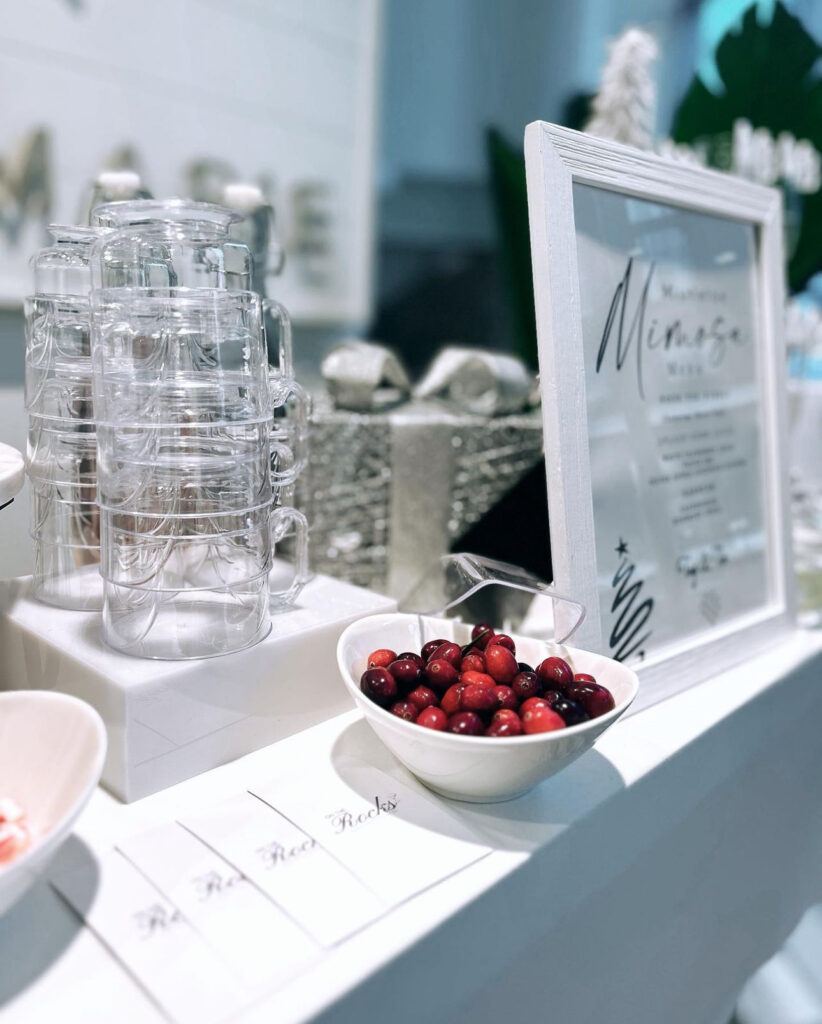 Stunning holiday corporate event mobile bartending bar setup in Madison, WI of white table, mimosa bar, shiny silver holiday décor, clear mug glasses stacked up on a white stand with a white bowl of red berries for garnishes next to a white framed mistletoe mimosa menu