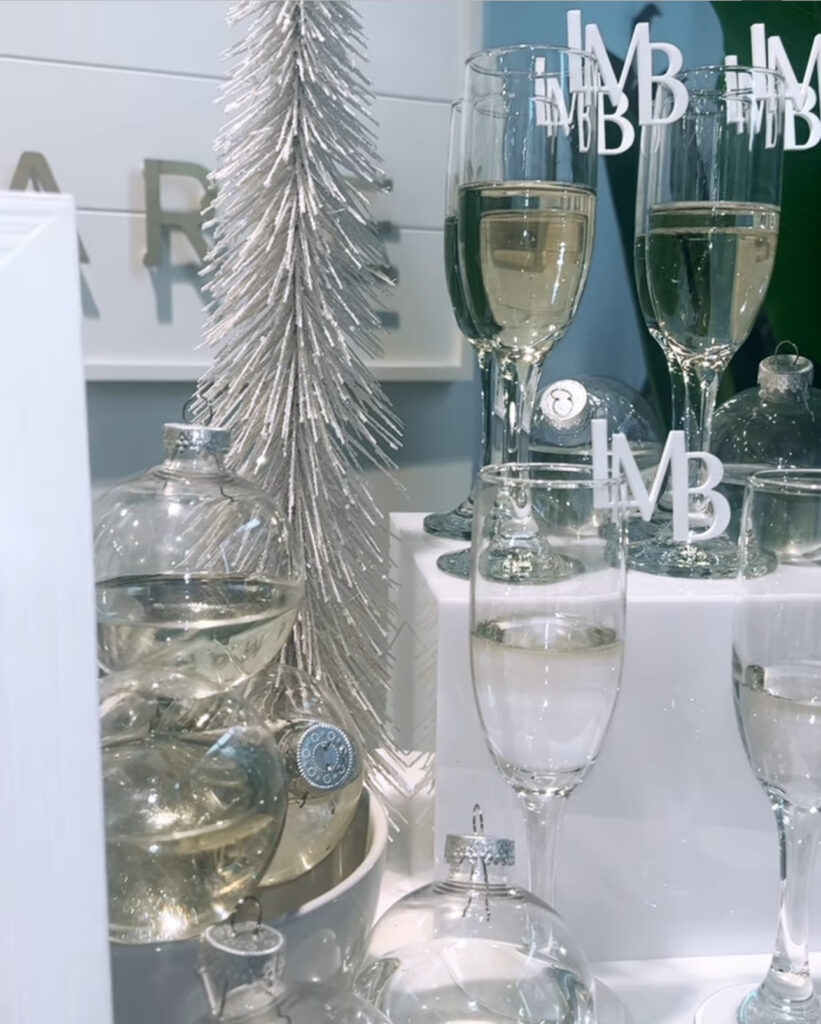 Stunning holiday corporate event mobile bartending bar setup in Madison, WI of white table, mimosa bar, shiny silver holiday décor, champagne glasses with drink stirrers of company logo initials and holiday clear ball ornaments filled with champagne for a gorgeous design touch