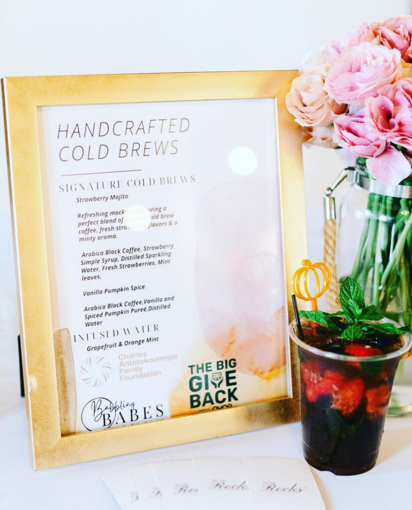 Closeup of handcrafted cold brew strawberry mojito mocktail with fresh mint leave garnish on top and a pumpkin drink stirrer and a clear jar of pink and blush roses and gold menu display setup by On The Rocks Mobile Bartending for a community event in Milwaukee WI