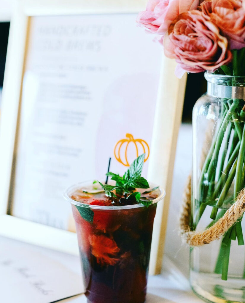 Closeup of handcrafted cold brew strawberry mojito mocktail with fresh mint leave garnish on top and a pumpkin drink stirrer and a clear jar of pink and blush roses and gold menu display setup by On The Rocks Mobile Bartending for a community event in Milwaukee WI