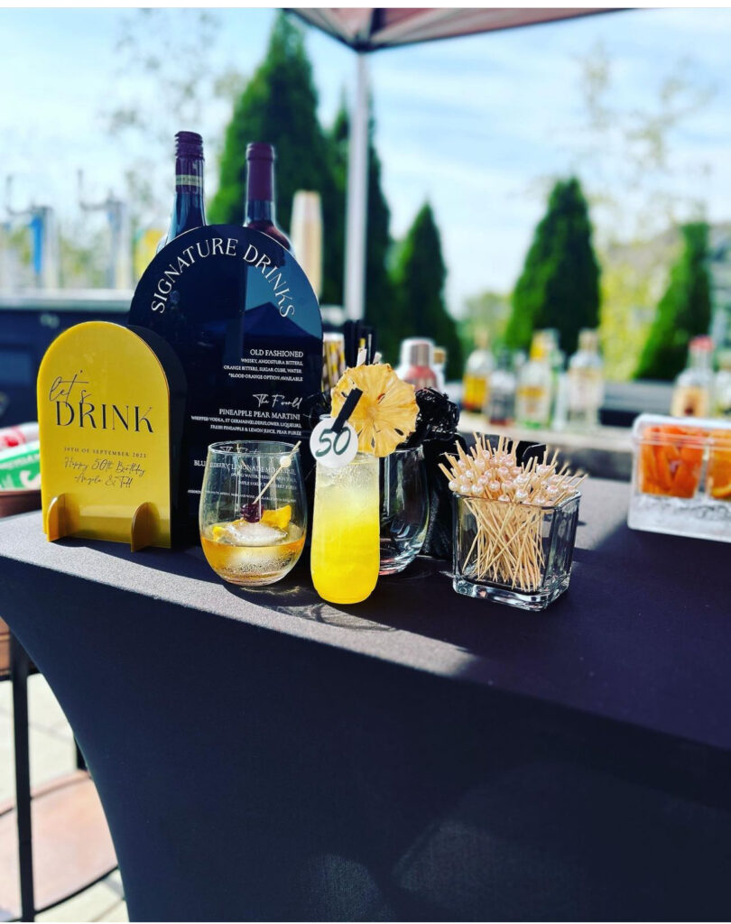 50th birthday event in Madison, WI bar setup by On the Rocks Mobile Bartending Company with acrylic rounded black drink menu and yellow event sign to match the event theme, an old fashioned with a pearl drink stirrer holding a cherry and fresh fruit garnishes as well as a vibrant refreshing yellow pineapple pear martini in a tall skinny clear glass with dried pineapple slice garnish and a pinned “50” drink topper attached to the rim. The table has a black tablecloth and is located under a tent outdoors.