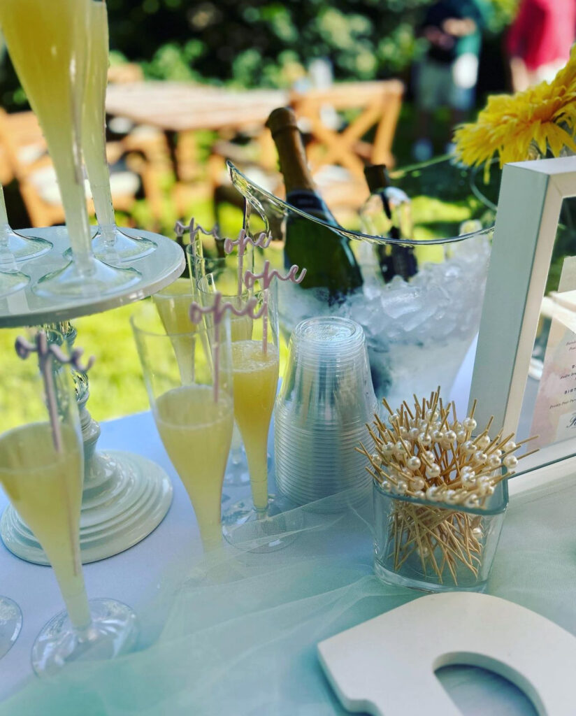 Display of a bar setup by On the Rocks Mobile Bartending Co for a Milwaukee event, first birthday party with a yellow, sunshine theme, tall clear glasses with pear puree and pineapple juice blended and pink drink stirrers with the word “one” written in cursive at the end sticking out of the drinks displayed on a white table and a beautiful cake stand as well. A small clear jar with pearl drink stirrers is also displayed for this outdoor backyard event