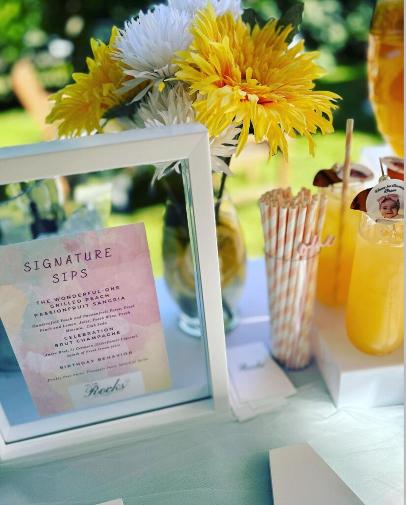 Display of a bar setup by On the Rocks Mobile Bartending Co for a Milwaukee event, first birthday party with a yellow, sunshine theme, grilled peach passionfruit sangria in a tall clear glass with fresh grilled peach garnish on a stick with a large pearl bead, pastel peach striped paper straws and a picture of the birthday girl as a drink topper, a white framed pastel watercolored menu, and a clear vase with white and yellow flowers and vibrant yellow lemon slices inside the vase