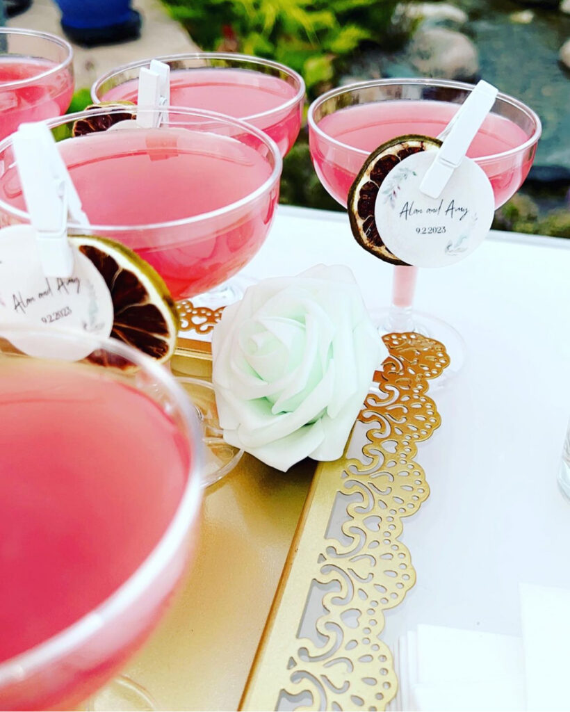 Display of beautiful custom bar setup by On the Rocks Mobile Bartending Co for a Milwaukee outdoor wedding of dark pink cosmos with dried fruit garnishes pinned to the rim along with a circular drink topper of the bride and grooms name and date and floral designs, lined up on a white table, some on a vintage ornate gold tray with a pastel green roses decorating the tray