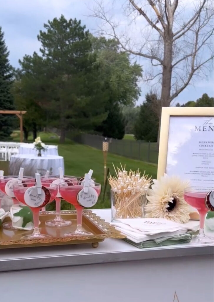 Display of custom bar setup by On the Rocks Mobile Bartending Co for a Milwaukee outdoor wedding of dark pink cosmos with dried fruit garnishes pinned to the rim along with a circular drink topper of the bride and grooms name and date and floral designs, lined up on a white table, some on a vintage ornate gold tray with a pastel green roses decorating the tray