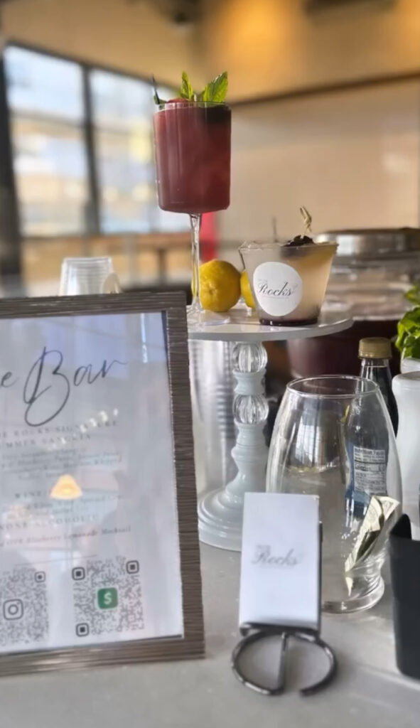 Display of custom bar setup by On the Rocks Mobile Bartending Co for a Milwaukee large indoor event with gorgeous tall cocktail glass with deep red drink and fresh mint leave and fruit garnishes and an elegant framed bar menu