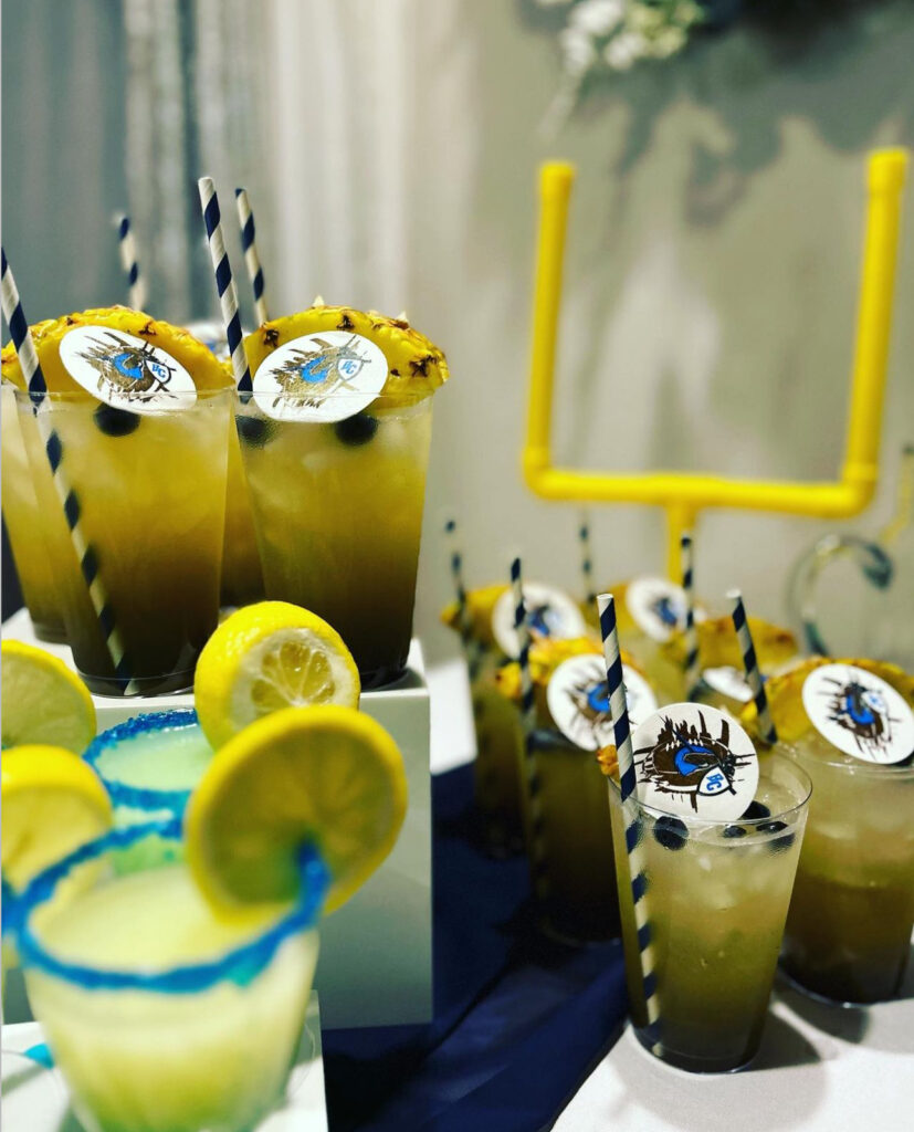 Bar setup of mocktail for end of season football team party event with different juice options, one in a small clear glass with blue salt along the rim to match the team colors and a fresh lemon slice on the rim; the other in a taller glass with fresh blueberries and pineapple slice garnishes, a blue and white striped straw and team logo drink topper. There is a small goalpost decoration behind the drinks for an added special detail for the event