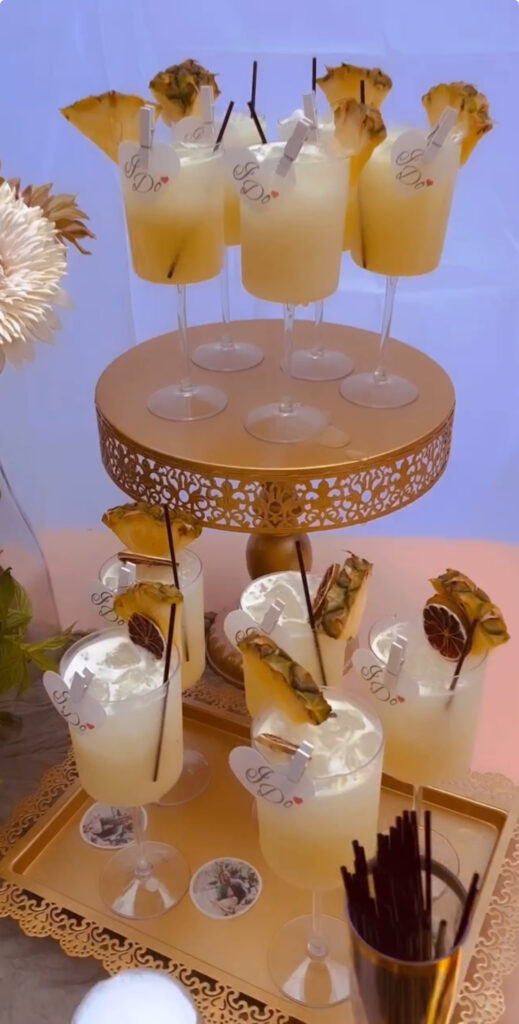 Gorgeous bar setup display designed and created by On the Rocks Mobile Bartending Company to fit the theme for an engagement party in Milwaukee, WI with a gold tray of yellow cocktails with fresh pineapple slice garnishes on some and dried fruit garnishes on others. They all have a paper heart pinned to the rim that says “I Do” in cursive with a red heart. On the drink display tray are paper circles with the couple’s engagement photos for an added level of special detail for the party