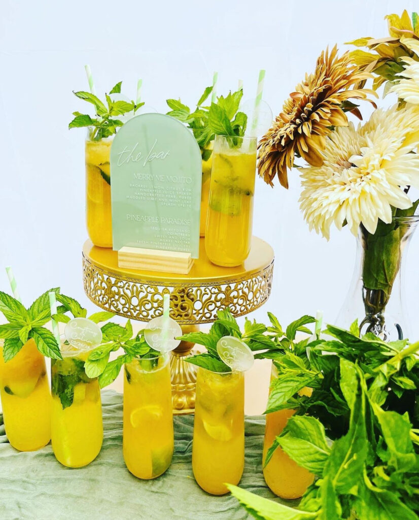 Cute bar setup display designed and created by On the Rocks Mobile Bartending Company to fit the theme for an engagement party in Milwaukee, WI. The drinks are setup on a table with a green tablecloth draped over it with a yellow mojito, fresh mint leave garnishes and a pink and white striped paper straw, some are displayed on a gold cake stand with a cute rounded acrylic green bar menu sign and a vase with white and gold flowers is behind it