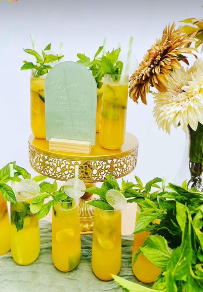 Cute bar setup display designed and created by On the Rocks Mobile Bartending Company to fit the theme for an engagement party in Milwaukee, WI. The drinks are setup on a table with a green tablecloth draped over it with a yellow mojito, fresh mint leave garnishes and a pink and white striped paper straw, some are displayed on a gold cake stand with a cute rounded acrylic green bar menu sign and a vase with white and gold flowers is behind it