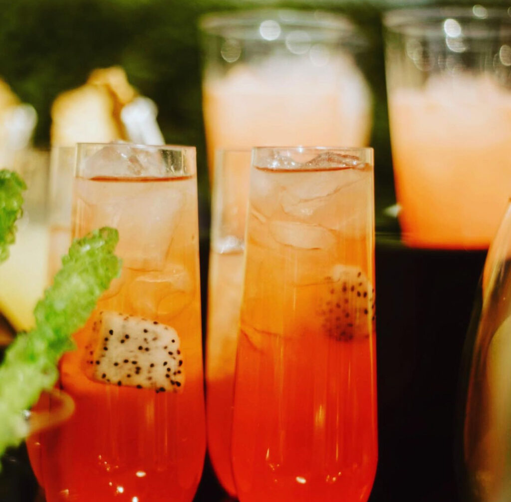 Gorgeous bar setup designed and created by On the Rocks Mobile Bartending Company for a Milwaukee event with a vintage, moody theme, black table and risers displaying tall vibrant red drinks with passionfruit cubes inside drink and a cup of green rock candy sticks offered next to the drinks