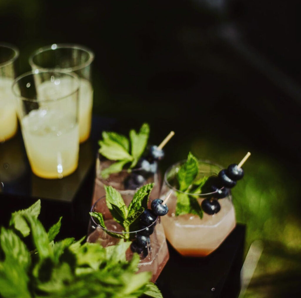 Gorgeous bar setup designed and created by On the Rocks Mobile Bartending Company for a Milwaukee event with a vintage, moody theme, black table and risers displaying drinks with fresh mint garnish as well as fresh blueberries on a stick resting inside glass