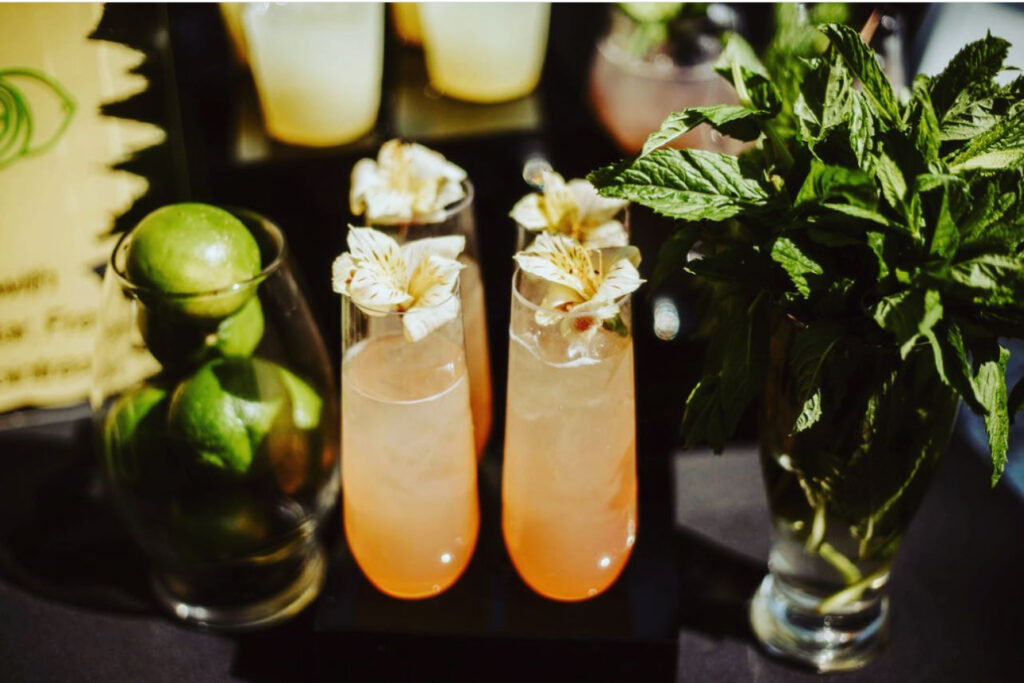 Beautiful peach colored drinks with floral garnishes for a custom bar setup on a black table with vibrant green limes in clear glass and green plants to add detail, amazing curated event bar setup by On the Rocks Mobile Bartending in Milwaukee, WI