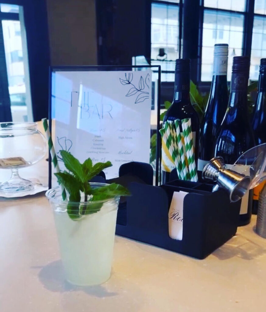 Bar setup by On the Rocks Mobile Bartending Company for a Milwaukee event with fresh lemonade and mint garnish and green and white striped paper straw, an elegant clear and thin black framed menu with black and white leaves is behind the drink and napkin/straw holder