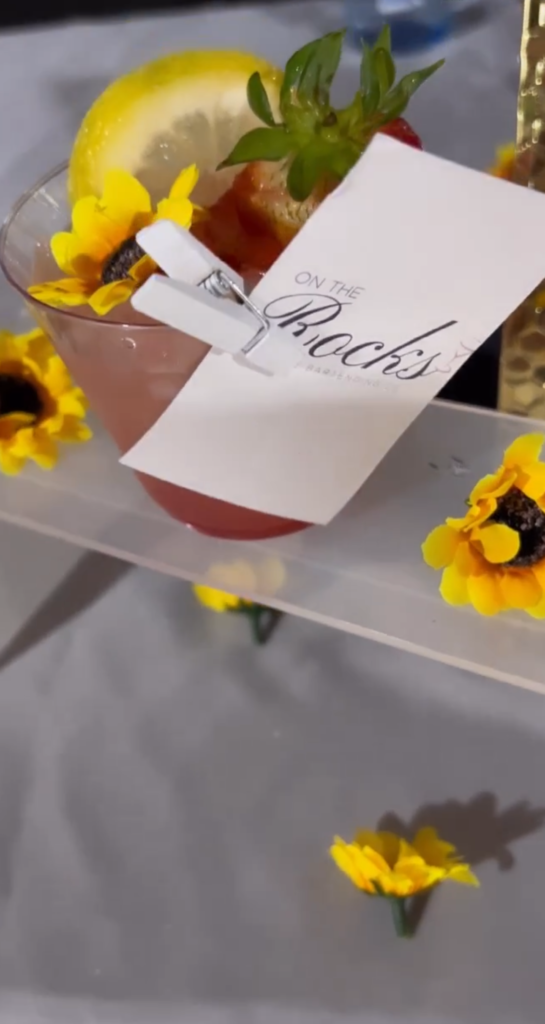 Display of a bar setup by On the Rocks Mobile Bartending Co for a Milwaukee event with a beautiful vibrant pink drink in a clear glass with fresh strawberry and lemon slice garnishes as well as a pretty yellow flower topping the drink. Pretty yellow flowers are decorating the bar display for extra wow factor making it a beautiful bar display matching the theme of the event