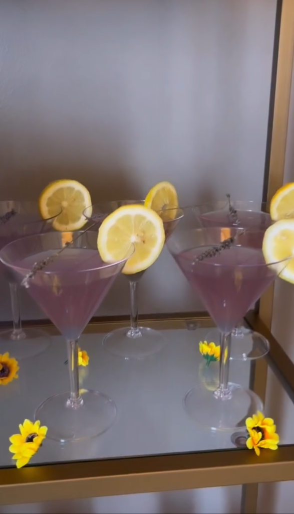 Stunning gold and clear vintage shelf bar setup display designed by On the Rocks Mobile Bartending Company for a Milwaukee community event, purple martinis with a sprig of lavender resting over the glass and a fresh lemon slice on the rim are lined up on the shelf with little yellow flowers decorating the shelf around the drinks for an added layer of detail making the look special