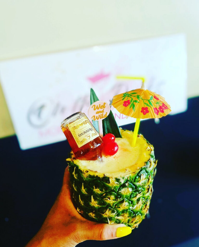 A fun tropical themed drink crated by On the Rocks Mobile Bartending of a pineapple colada inside of a hollowed out pineapple topped with a tropical umbrella straw, cherries, pineapple leaves, a mini bottle of Hennessy tipped over and a drink stirrer stick that says “wet and wild”