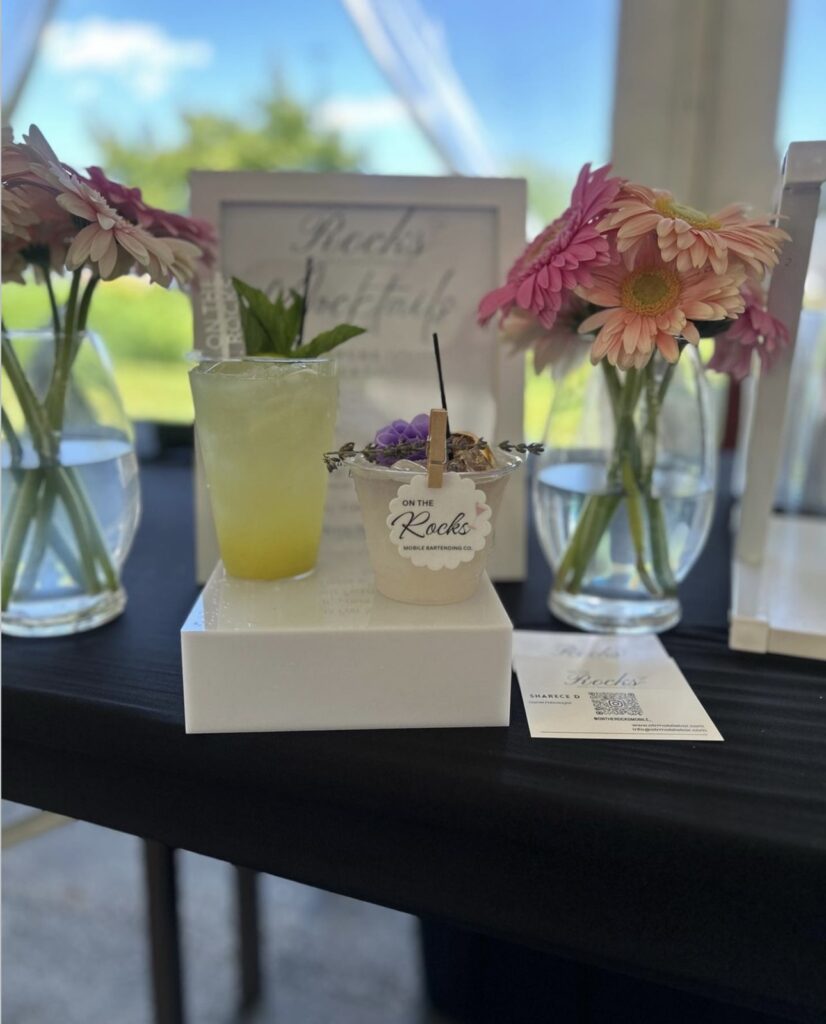 Classy event bar setup with 2 cocktails, a high top yellow drink with fresh fruit slice and mint garnishes and a short glass with beautiful purple flower garnish, lavender sprig and dried fruit garnish with On The Rocks Mobile Bartending logo pinned to the rim displayed on a white riser with pretty pink and peach daisies in vases on either side