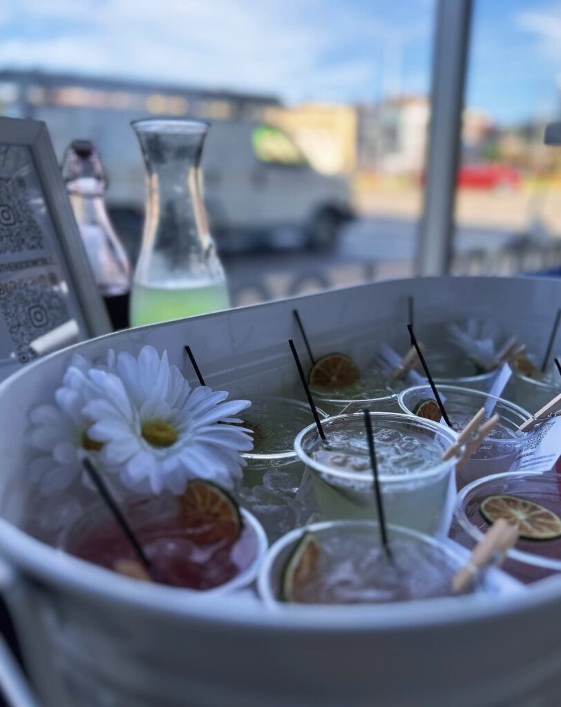 Pretty private event bar setup with vibrant pink and yellow cocktails and mocktails and dried fruit garnishes displayed in a white oval bin and white daisies sprinkled in between the drinks