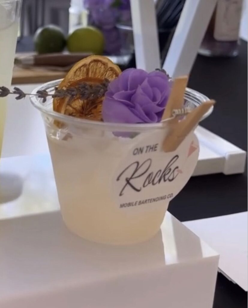 Pretty cocktail with beautiful purple flower garnish, lavender sprig and dried fruit garnish with On The Rocks Mobile Bartending logo pinned to the rim displayed on a white riser