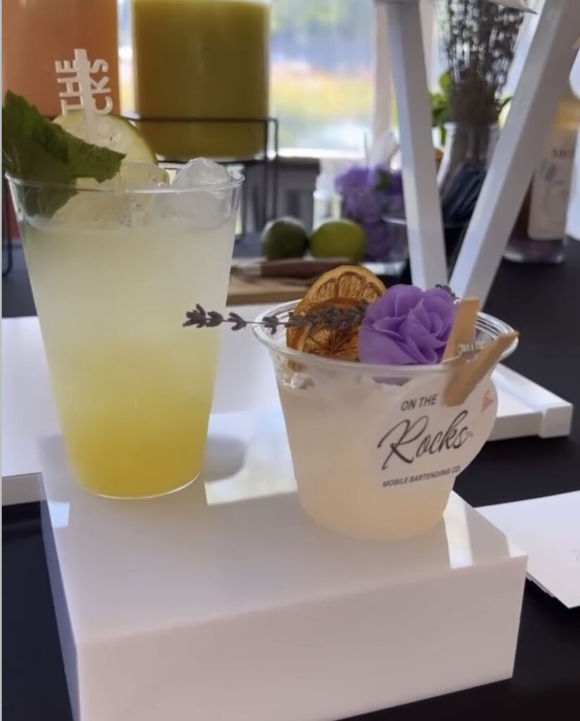 Classy event bar setup with 2 cocktails, a high top yellow drink with fresh fruit slice and mint garnishes and a short glass with beautiful purple flower garnish, lavender sprig and dried fruit garnish with On The Rocks Mobile Bartending logo pinned to the rim displayed on a white riser