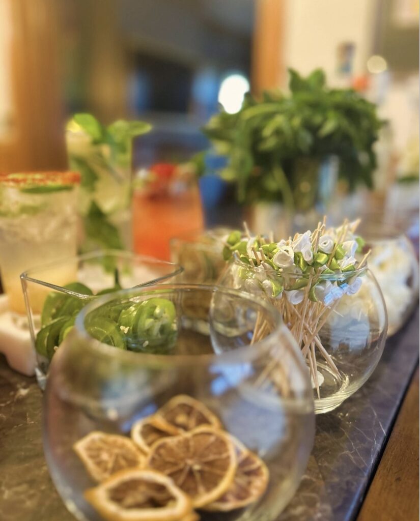 Cocktail garnishments of dried fruit slices, jalapenos and fabric flower wooden stir sticks in classy glass bowls for private event mobile bartending display