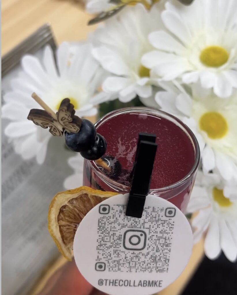 Gorgeous fresh Blueberry Elderflower French 75 cocktail with dried fruit garnish pinned to rim and wooden butterfly stir stick with fresh blueberries created for a bar setup by On The Rocks Mobile Bartending for a Milwaukee women in business networking event, drink is shown closeup with white daisies as a backdrop