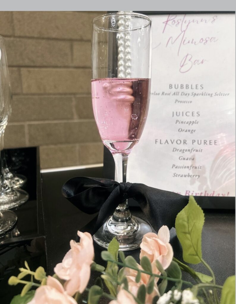 Rosé with black fabric bow tied around stem and pink roses displayed in front of it as a special private event mimosa bar setup by On The Rocks Mobile Bar in Milwaukee