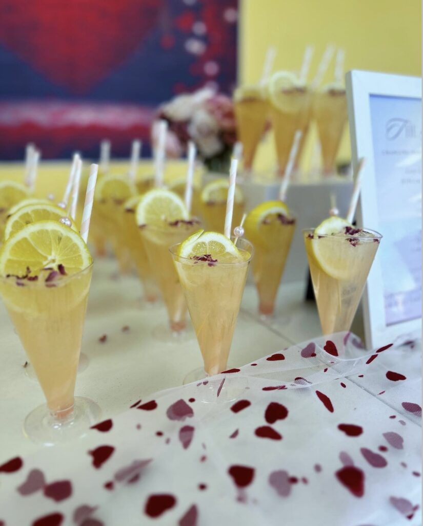 Cute bar setup display for youth girl private event mocktails fancy Hibiscus Rose Spritzers with fresh lemon slice garnishes, rose petals sprinkled on top, a pink pearl stir stick and pastel pink and white striped paper straws with heart confetti and toile lining the table. Event bartending design made special for the youth event by On The Rocks Mobile Bartending co in Milwaukee WI